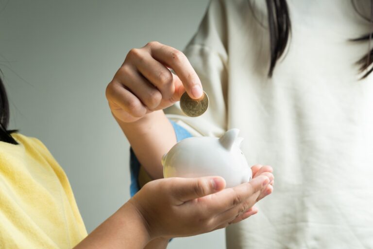 Cropped image of a child's hands holding a piggy bank and the mother's hand putting a coin; concept of 'how to save money when moving to Weymouth'.